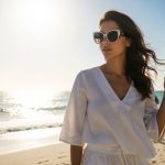 A woman wearing stylish cheap designer sunglasses on a sunny beach, dressed in a white summer outfit