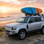 SUV parked by beach with surfboard secured on roof rack at sunrise